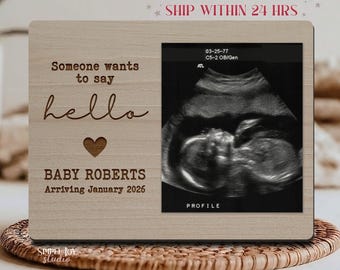 May include: A wooden picture frame with the text "Someone wants to say hello" and "BABY ROBERTS Arriving January 2026" engraved on it. The frame holds a sonogram image. The frame is light brown.