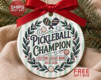 May include: A round ceramic ornament with a red satin ribbon bow. The surface features light green vertical stripes, a wreath of green leaves with red berries, two pickleball paddles, three balls, and the text "PICKLEBALL CHAMPION".