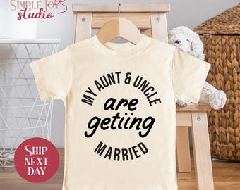 May include: Cream-colored toddler t-shirt with the text "MY AUNT & UNCLE are getting MARRIED" in black. The shirt is hanging on a wooden hanger. A stuffed animal and a small wooden stool are in the background.
