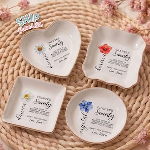 May include: Four white ceramic trinket dishes in heart, square, and round shapes. Each dish features a floral illustration, the text Chapter Seventy, a name, and a 70th birthday message. The dishes have gold-toned rims and personalized inscriptions.