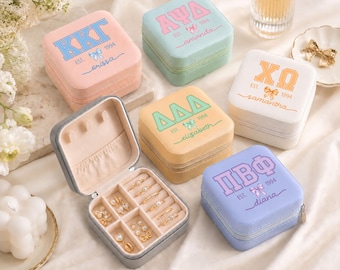 May include: A collection of square, zippered jewelry cases in pastel colors, each featuring Greek letters, a small bow icon, the text 'EST. 1994', and a personalized name. One case is open, revealing internal compartments holding gold rings.