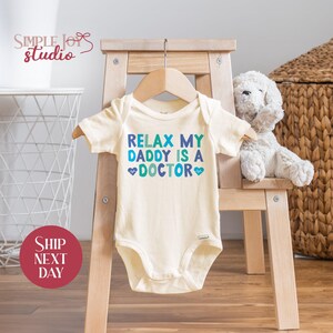 May include: A white baby onesie with a blue and green graphic that says "Relax My Daddy Is A Doctor" with two heart symbols.