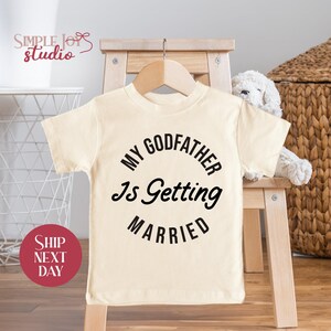 May include: Cream-colored toddler t-shirt with the text "MY GODFATHER Is Getting MARRIED" in black. The shirt is hanging on a wooden hanger. The Simple Joys Studio logo is visible in the upper left corner.