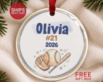 May include: A round white ceramic ornament with a red ribbon hanger. The design features the name Olivia in blue, the number #21 in tan, and the year 2026 in blue. Below the text is a baseball, a leather glove, and a wooden bat illustration.