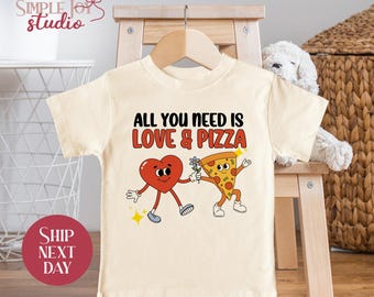 May include: A beige t-shirt with a graphic of a heart and a pizza slice holding hands. The text "All you need is love & pizza" is printed on the shirt.