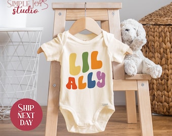 May include: A cream colored baby onesie with the text "Lil Ally" in rainbow colored letters. The onesie is hanging on a wooden hanger.