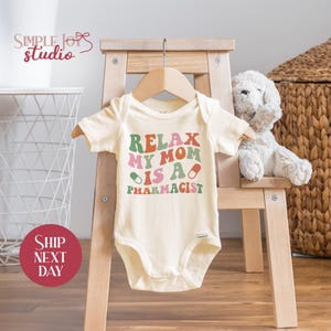 May include: A white baby onesie with the text "Relax My Mom Is A Pharmacist" in colorful, retro-style lettering. The text is surrounded by two green and red pill illustrations.