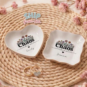 May include: Two white ceramic ring dishes with gold trim, one heart-shaped and one square-shaped, each featuring a floral design and the text "Chaos coordinator." The heart-shaped dish has the name "Emily" and the square-shaped dish has the name "Joanna." Two gold rings with a heart-shaped diamond are also visible.