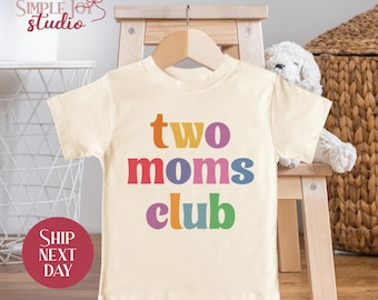May include: A cream colored t-shirt with the text "two moms club" in rainbow colors. The shirt is hanging on a wooden hanger.
