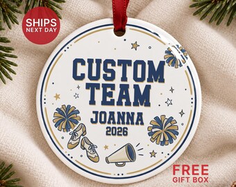 May include: A round white ceramic ornament with a red ribbon hanger. The design features dark blue text reading CUSTOM TEAM, JOANNA, and 2026, surrounded by illustrations of two pom-poms, a megaphone, and two dance shoes with gold stars.