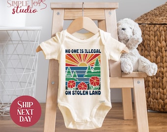 May include: Cream-colored baby bodysuit with a graphic design featuring a sunset, trees, and flowers. The text "NO ONE IS ILLEGAL ON STOLEN LAND" is printed on the front. The bodysuit is hanging on a wooden hanger.