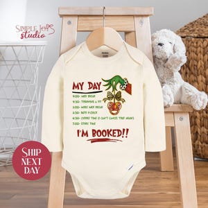 May include: Cream-colored baby onesie with long sleeves, featuring the text "MY DAY" and a schedule of activities. The design includes a Grinch-themed graphic with a pacifier and bow. The words "I'M BOOKED!!" are also printed on the onesie.