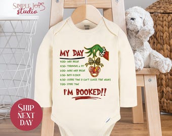 May include: Cream-colored baby onesie with long sleeves, featuring the text "MY DAY" and a schedule of activities. The design includes a Grinch-themed graphic with a pacifier and bow. The words "I'M BOOKED!!" are also printed on the onesie.