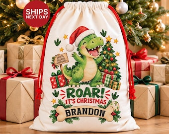 May include: A cream-colored drawstring gift sack with red cords featuring a green dinosaur wearing a Santa hat, a Christmas tree, and wrapped presents. The text reads: SANTA SAURUS APPROVED, ROAR! IT'S CHRISTMAS, and the name BRANDON on a bone.