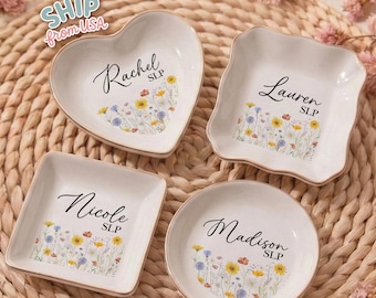May include: Four personalized ceramic trinket dishes in heart, square, round, and scalloped square shapes. Each dish is white with a floral design and a name in elegant script. The dishes have a gold rim.