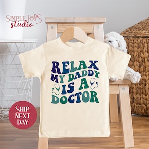 May include: A cream-colored baby t-shirt with the text "Relax My Daddy Is A Doctor" in a colorful retro font. The text is surrounded by two blue and green stethoscope graphics.