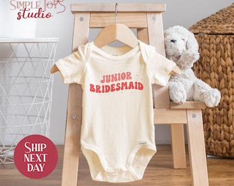 May include: Cream-colored baby bodysuit with the words "Junior Bridesmaid" in red script. The short-sleeved onesie hangs on a wooden hanger, displayed on a small wooden chair. A stuffed white dog sits nearby. The Simple Joys Studio logo is visible.