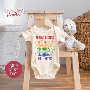 May include: A white baby onesie with a rainbow-colored possum with wings and the text "Trans Rights or I Bites".