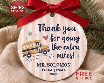 May include: A round ceramic ornament with a red satin ribbon, featuring a pink and white striped background. It displays a yellow school bus graphic, a small heart, stars, and the text: Thank you for going the extra miles! MR. SOLOMON FROM. JESSIE 2026.