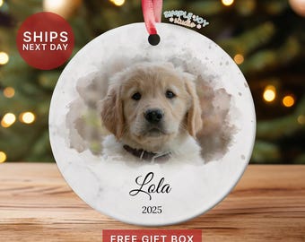 May include: A round, white ceramic ornament featuring a watercolor image of a golden retriever puppy. The puppy's name, "Lola," and the year "2025" are written in script. A red ribbon is attached for hanging.