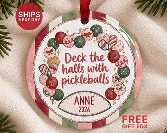 May include: A round ceramic Christmas ornament with a red ribbon hanger. The design features a wreath of pickleballs and paddles surrounding the text Deck the halls with pickleballs. Below, a label displays the name ANNE and the year 2026.