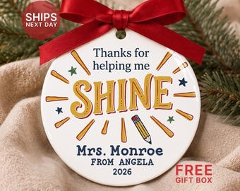 May include: A round white ceramic ornament with a red ribbon bow at the top. The text reads "Thanks for helping me SHINE" in blue and gold, with a pencil graphic, "Mrs. Monroe", "FROM ANGELA", and "2026" printed in dark blue.