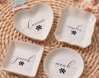 May include: Four white ceramic dishes in heart, square, round, and scalloped square shapes. Each dish has a gold rim, a name in cursive, and a paw print. The text "Lauren", "Sophia", "Jacob", and "Noah" are visible.