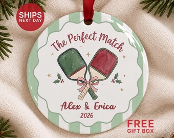 May include: A round ceramic ornament with a red ribbon hanger, featuring a green and red pickleball paddle illustration, the text The Perfect Match, Alex & Erica, and the year 2026, set against a light green and white striped background.