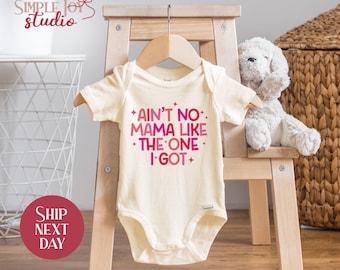 May include: Cream-colored baby onesie with the text "Ain't No Mama Like The One I Got" in pink and magenta. The onesie is displayed on a wooden hanger and a small wooden chair. A stuffed animal is to the right.