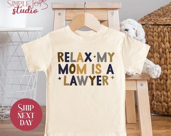 May include: Cream-colored toddler t-shirt with the text "RELAX MY MOM IS A LAWYER" in navy, brown, and gray lettering. The shirt is displayed on a wooden hanger and a small wooden chair.