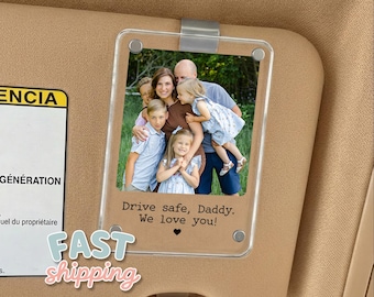 May include: A clear acrylic car visor photo holder with a family photo. The photo holder is attached to a tan car visor. The photo shows a family of five. Text on the photo holder reads "Drive safe, Daddy. We love you!"