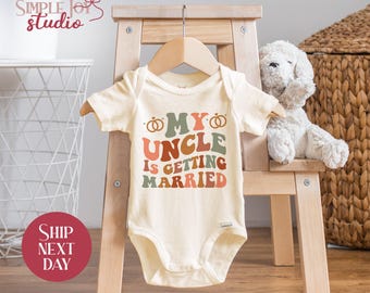 May include: Cream-colored baby onesie with the text "MY UNCLE IS GETTING MARRIED" in retro-style lettering. The design includes two wedding rings. The onesie is hanging on a wooden hanger on a small wooden chair.
