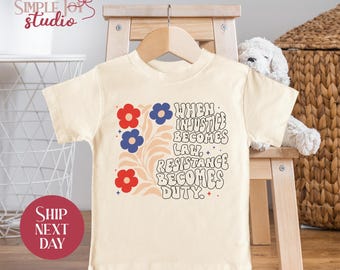 May include: Cream-colored t-shirt with the text "When Injustice Becomes Law, Resistance Becomes Duty." The shirt features red and blue flower graphics and a floral design. The shirt is displayed on a wooden hanger.