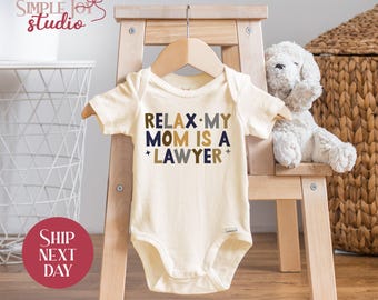 May include: A cream-colored baby onesie with the text "RELAX MY MOM IS A LAWYER" in navy blue and gold lettering.