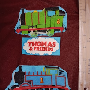 Puede incluir: Tres parches de tela con personajes de Thomas & Friends. Un parche muestra un tren verde con el número 6, otro muestra un tren azul con el número 1, y un tercer parche tiene el texto "THOMAS & FRIENDS" en rojo y blanco.