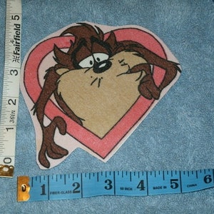 May include: A pink heart-shaped patch with a cartoon image of Taz the Tasmanian Devil inside. Taz is brown and has a mischievous expression on his face.