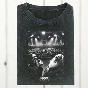 May include: Dark grey t-shirt with a black and white graphic depicting two people singing into microphones before a large audience. The image is set in a large hall with arched ceilings and bright lights.
