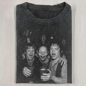 May include: Dark gray t-shirt featuring a black and white image of characters from The Lord of the Rings, including Gollum, holding mugs and cheering. The shirt is folded and has a crew neck.