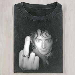 Op de afbeelding: Zwart T-shirt met een zwart-wit afbeelding van een persoon die de middelvinger opsteekt. De persoon draagt een ring. Het T-shirt is op een licht houten oppervlak gevouwen.