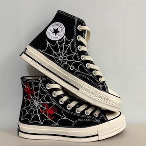 May include: Black high-top Converse sneakers with a white spiderweb and red spider design. The shoes have a white rubber sole and a black canvas upper.