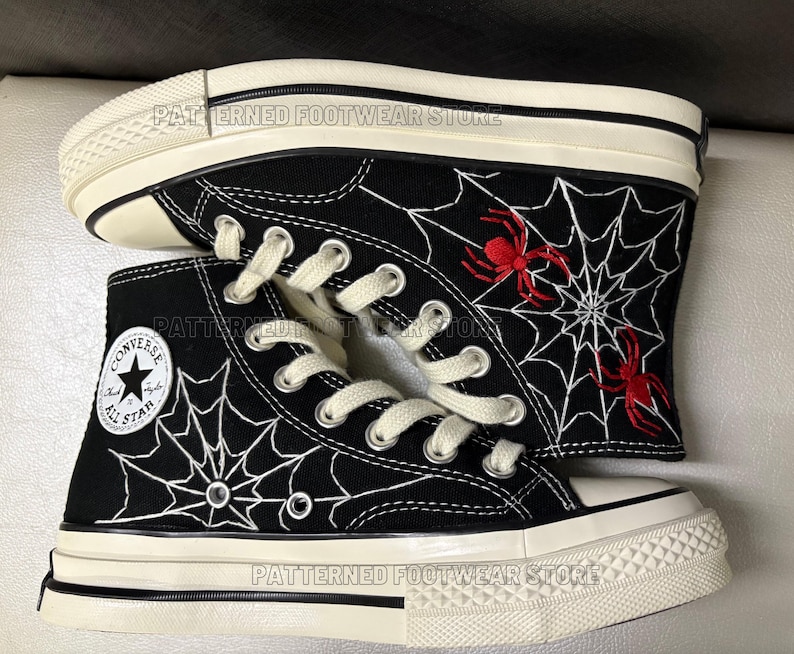 May include: Black high-top Converse sneakers with white spiderweb and red spider embroidery. The shoes have a white rubber sole and a black canvas upper. The Converse All Star logo is visible on the side of the shoe.