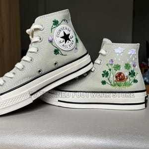 May include: A pair of light green high-top Converse trainers with embroidered four-leaf clovers, a snail, and raindrops. The trainers have white soles and black stitching.