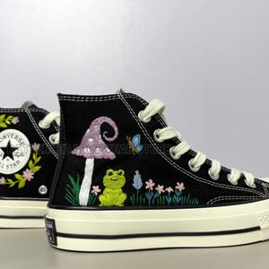 Op de afbeelding: Zwarte hoge sneakers met geborduurde ontwerpen. Eén schoen heeft een paddenstoel, kikker en vlinder, de andere een bloemenkrans rond een sterembleem. Witte veters en zolen.