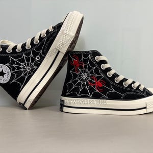 May include: Black high-top Converse sneakers with a white spiderweb design and red spiders. The shoes have a white rubber sole and the Converse logo is visible on the side.