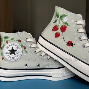 Könnte beinhalten: Ein Paar hellgrüne Converse-Sneaker mit hohem Schaft, bestickt mit Erdbeeren, Blättern und einem Marienkäfer. Das Converse All Star Logo ist an der Seite des Schuhs sichtbar.
