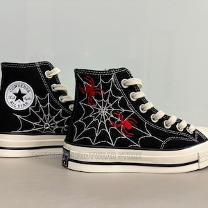 May include: Black high-top Converse sneakers with a white spiderweb and red spider embroidery. The shoes have a white rubber sole and a black star logo on the side.