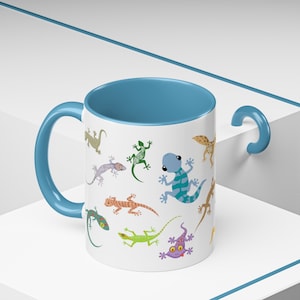 May include: A white ceramic mug with a blue rim and handle features a colorful pattern of cartoon lizards. The mug is filled with a light blue liquid.