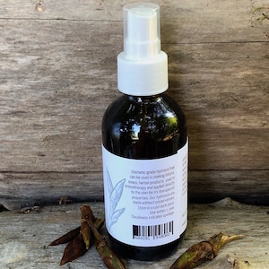 May include: A clear glass bottle with a white spray top filled with a dark liquid. The bottle has a white label with black text that reads "Cosmetic grade hydrosol that can be used in making lotions, soaps, herbal products, used in aromatherapy and applied directly to the skin for its therapeutic properties. Our hydrosols are made without preservatives. Store in a cool dark place. Use within 1 year. Cloudiness indicates spoilage." The bottle is sitting on a wooden surface with a few brown twigs.