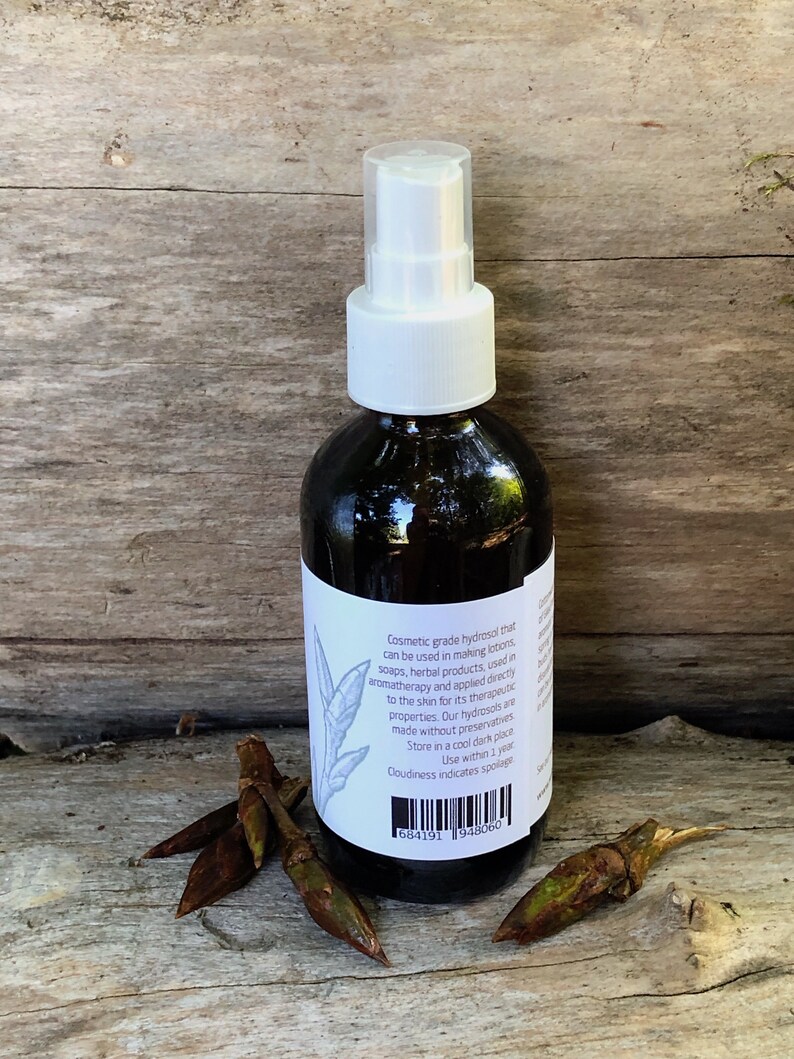 May include: A clear glass bottle with a white spray top filled with a dark liquid. The bottle has a white label with black text that reads "Cosmetic grade hydrosol that can be used in making lotions, soaps, herbal products, used in aromatherapy and applied directly to the skin for its therapeutic properties. Our hydrosols are made without preservatives. Store in a cool dark place. Use within 1 year. Cloudiness indicates spoilage." The bottle is sitting on a wooden surface with a few brown twigs.