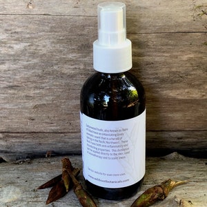 May include: A brown glass bottle with a white spray top filled with a clear liquid. The bottle has a white label with text that reads "Cedarwood buds, also known as Balm of Gilead have an intoxicating lovely aromatic scent that is a herald of spring in the Pacific Northwest. These buds have both anti-inflammatory and disinfecting properties. This distillation can be applied directly to the skin, used in aromatherapy and to scent linens. See our website for even more uses www.wildrootbotanicals.com"